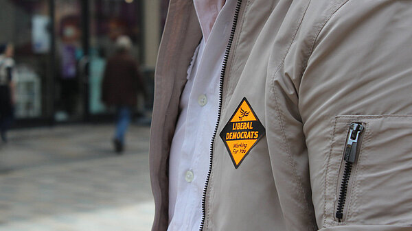 shot of person wearing grey jacket with orange lib dem sticker